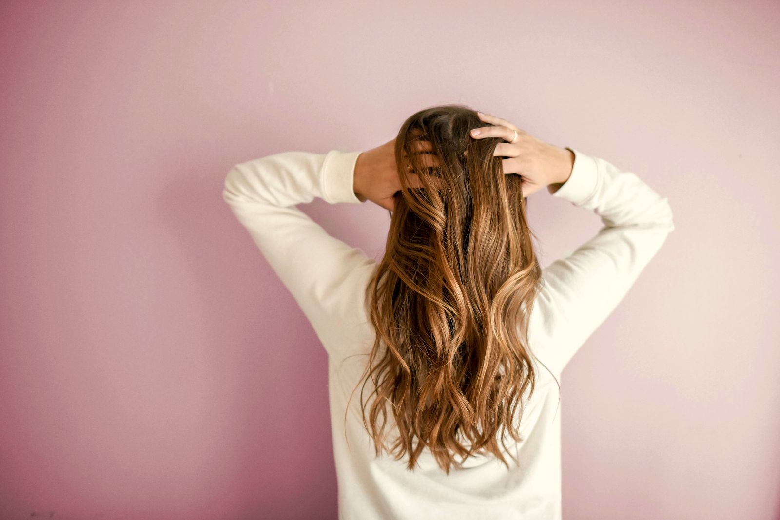 A woman seen from behind, hands in her long waves of brown hair painted with caramel balayage